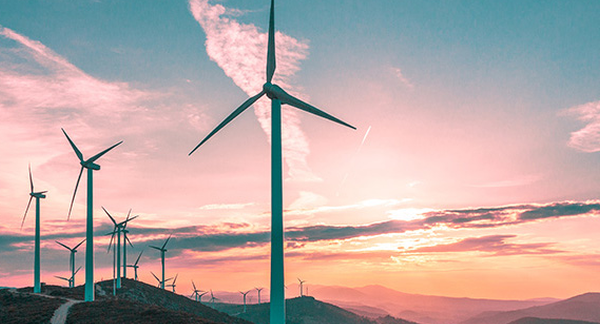 The image shows a row of wind turbines on a hilly terrain during what appears to be either sunrise or sunset, given the pink and orange hues in the sky. The turbines are positioned prominently against the sky, with their blades in various positions. This image captures renewable energy technology within a natural landscape, highlighting a harmonious blend of human innovation and the environment.