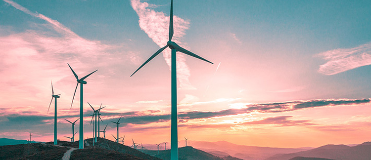 The image shows a row of wind turbines on a hilly terrain during what appears to be either sunrise or sunset, given the pink and orange hues in the sky. The turbines are positioned prominently against the sky, with their blades in various positions. This image captures renewable energy technology within a natural landscape, highlighting a harmonious blend of human innovation and the environment.