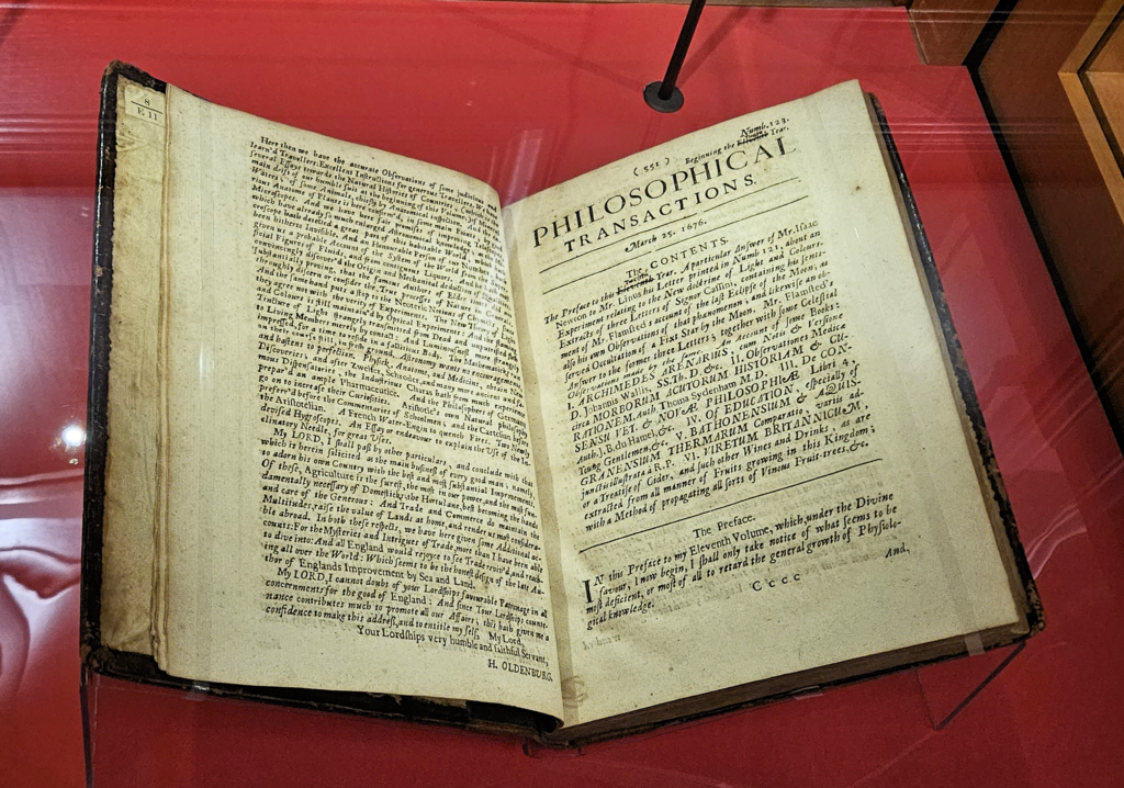 The image depicts an open antique book displayed under glass. The title on the right page reads “PHILOSOPHICAL TRANSACTIONS” and is surrounded by a decorative border. The left page contains dense, printed English text.
