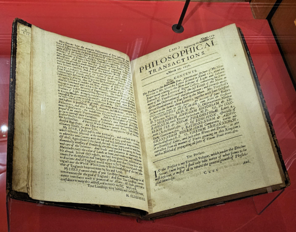 The image depicts an open antique book displayed under glass. The title on the right page reads “PHILOSOPHICAL TRANSACTIONS” and is surrounded by a decorative border. The left page contains dense, printed English text.