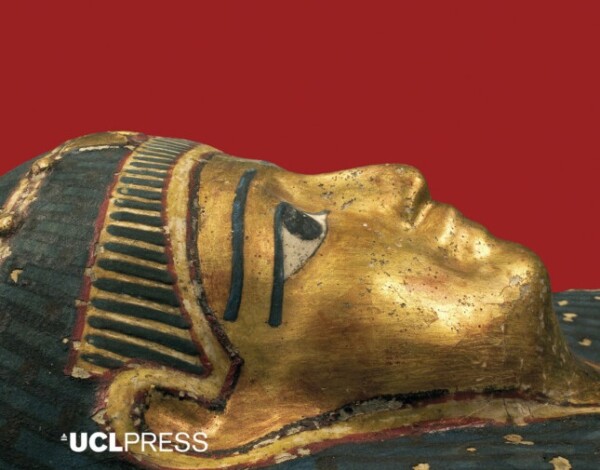 Close-up of an ancient Egyptian artifact depicting a golden sphinx with intricate detailing in black and red against a red background. The sphinx has a human head with pharaonic headcloth and a lion’s body