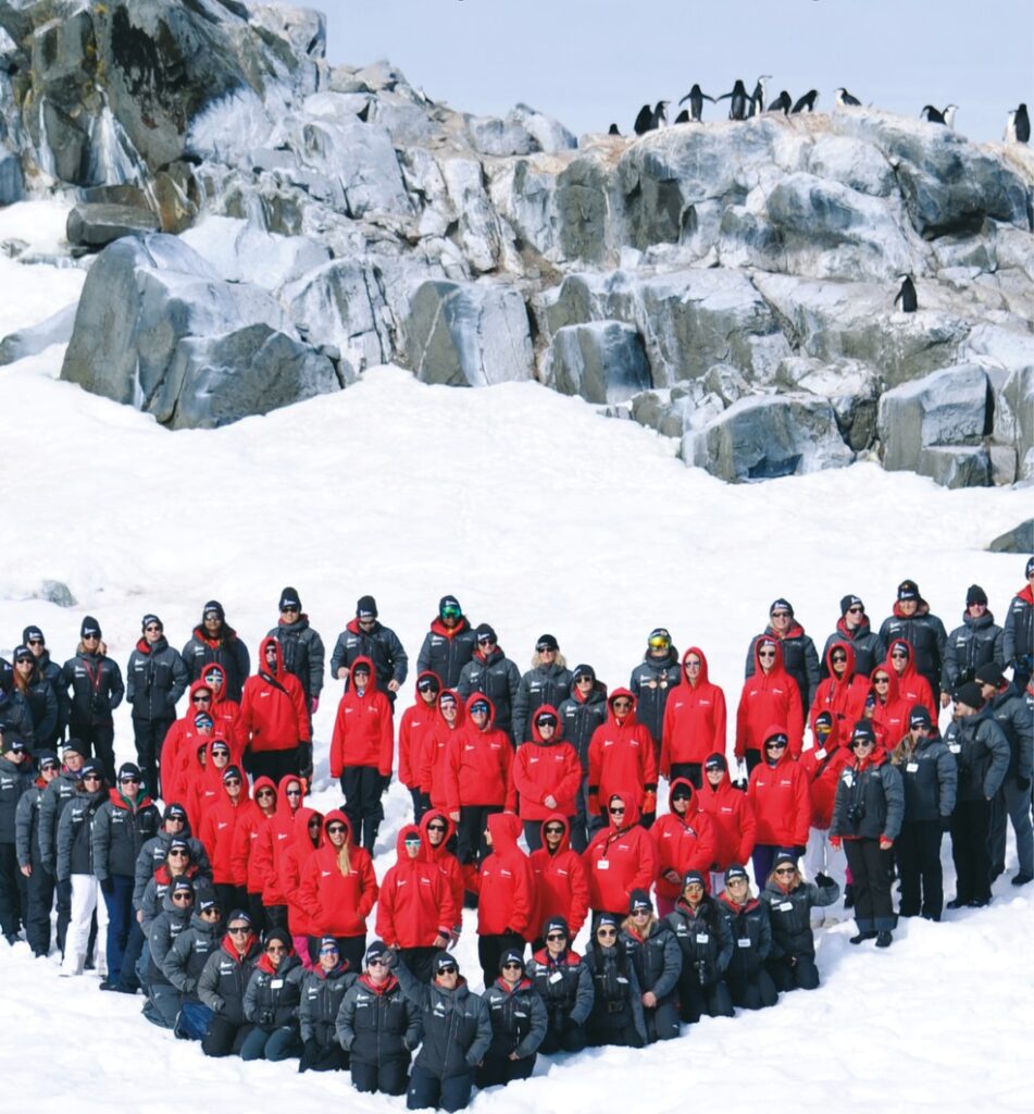 The image depicts a large group of individuals wearing red and black parkas in a heart formation. Those in red parkas are at the centre, and those in black are on the edges. They are gathered on a snowy landscape in front of a rocky area with penguins in the background.