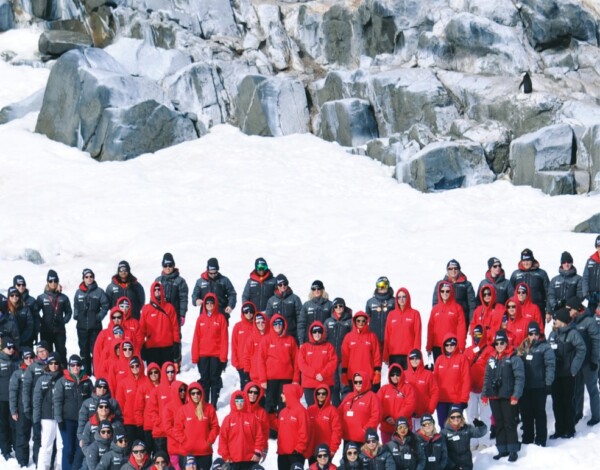 The image depicts a large group of individuals wearing red and black parkas in a heart formation. Those in red parkas are at the centre, and those in black are on the edges. They are gathered on a snowy landscape in front of a rocky area with penguins in the background.