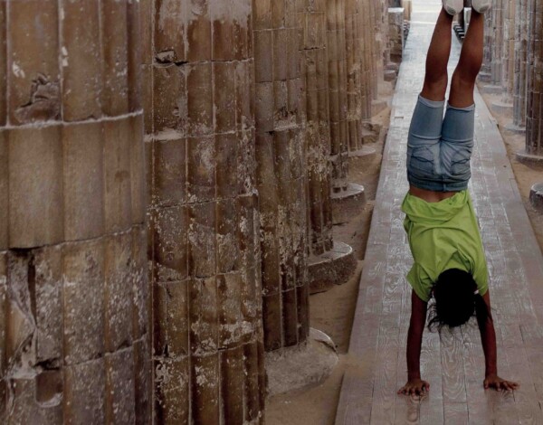 A person in a green shirt and denim shorts performs a handstand between ancient stone columns.