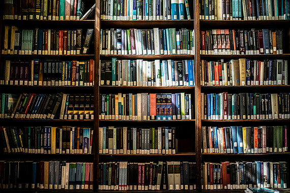 A large library bookshelf filled with books of various sizes and colors. The books are tightly packed, and the shelf appears to be well-organized, with some books standing upright and others stacked horizontally. The lighting casts a warm glow on the spines of the books