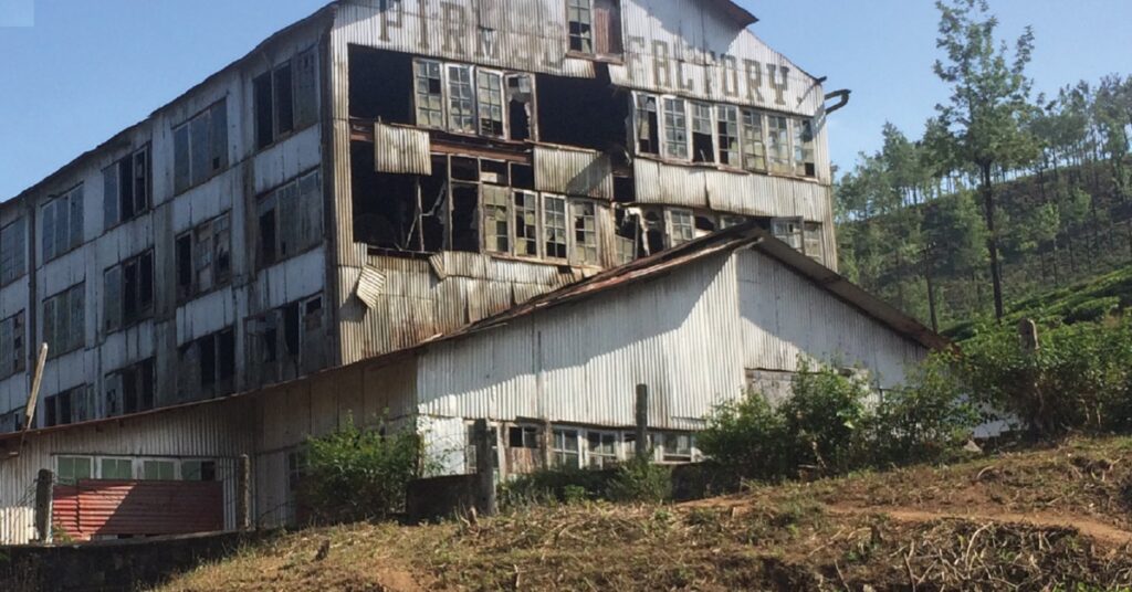 The image shows a large, dilapidated industrial building with multiple levels, surrounded by vegetation. The structure appears to be abandoned and in a state of disrepair, with broken windows and rusted metal. The word “FACTORY” is visible on the top of the building