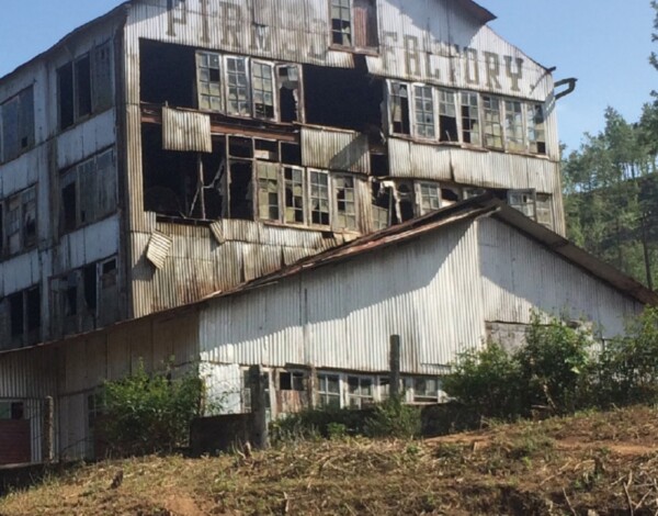 The image shows a large, dilapidated industrial building with multiple levels, surrounded by vegetation. The structure appears to be abandoned and in a state of disrepair, with broken windows and rusted metal. The word “FACTORY” is visible on the top of the building
