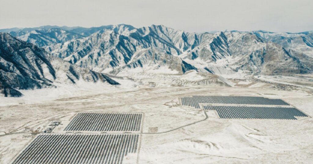 Aerial view of vast solar panel fields in a snowy landscape with rugged mountains in the background.