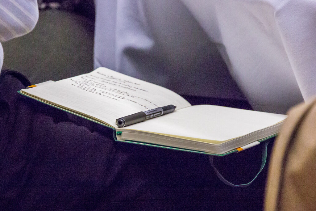 A notebook with a black pen resting on it sits open on a dark blue surface. The notebook is mostly empty with handwritten notes in the top left corner.