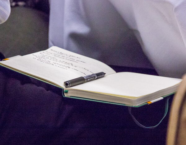 A notebook with a black pen resting on it sits open on a dark blue surface. The notebook is mostly empty with handwritten notes in the top left corner.