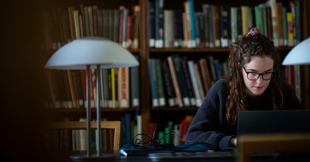 Individual studying on a laptop in a library, surrounded by books and under the illumination of desk lamps.