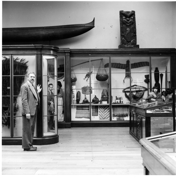 A man at the British Museum ethnography gallery is standing beside a glass display case containing various cultural artefacts. Another glass display case lines the wall behind, containing woven baskets, pottery and textiles. A wooden canoe and a carved wooden artefact are placed above the case.