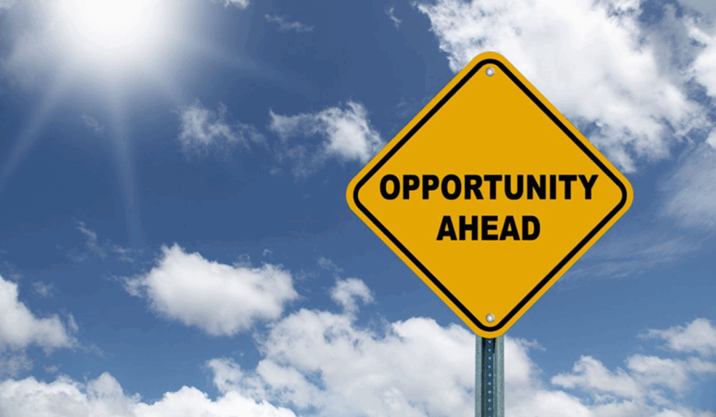 A yellow diamond-shaped road sign saying "OPPORTUNITY AHEAD" against a blue sky with clouds.