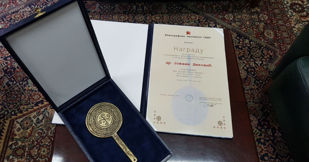 An open presentation box containing a circular gold-toned award with an ornate design, placed on a dark blue velvet lining, next to an open folder holding a certificate written in cyrrilic script. Both items are displayed on a wooden table with a patterned rug in the background.