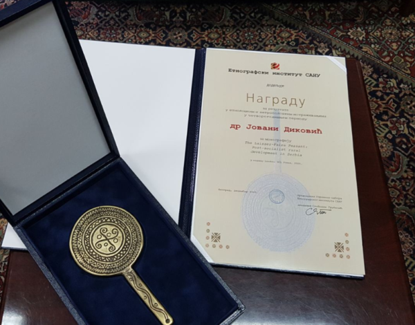 An open presentation box containing a circular gold-toned award with an ornate design, placed on a dark blue velvet lining, next to an open folder holding a certificate written in cyrrilic script. Both items are displayed on a wooden table with a patterned rug in the background.