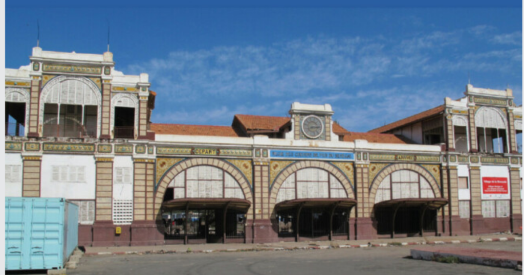 Exterior view of the Terminus Market in Dakar, showcasing ornamental arches, a clock, and a light blue shipping container in front.