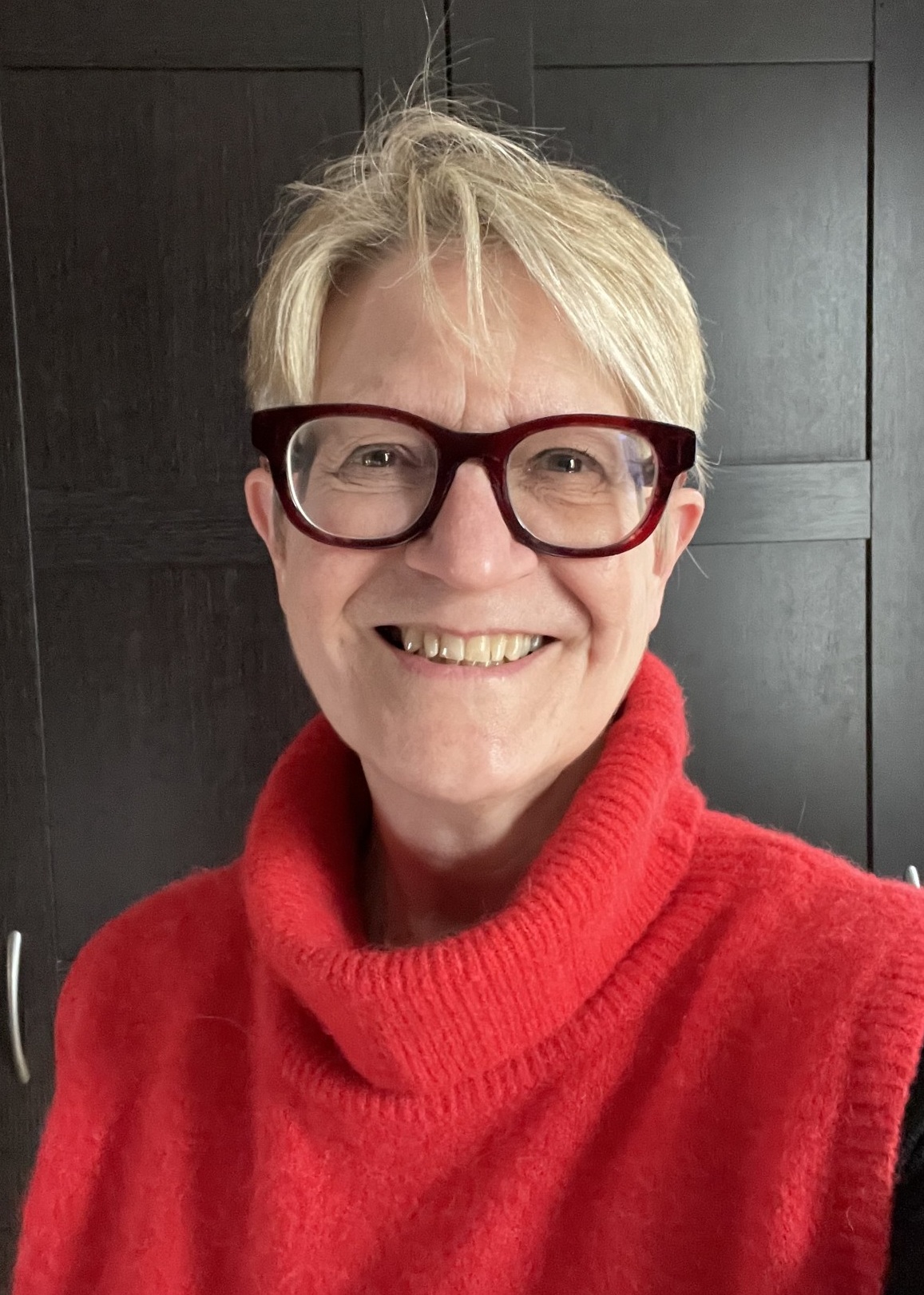 A portrait of a smiling woman with short blonde hair. She is wearing thick, dark-framed glasses and a vibrant red roll-neck jumper. She is positioned in front of a dark, panelled door background.