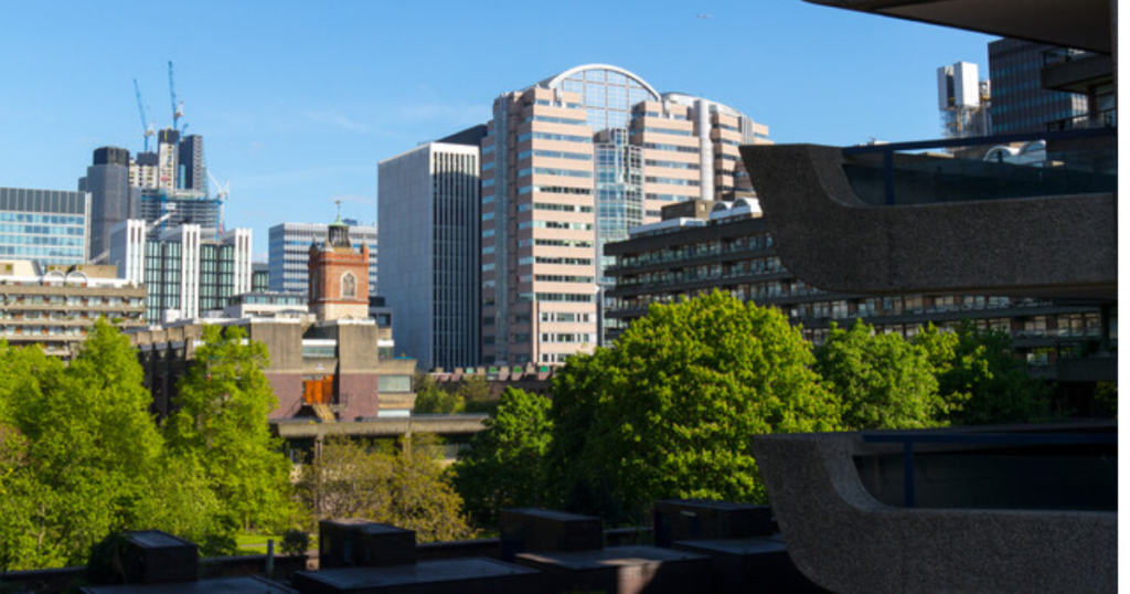 London skyline viewed from the Barbican, with modern office towers, leafy trees, and dark angular Barbican buildings in the foreground.