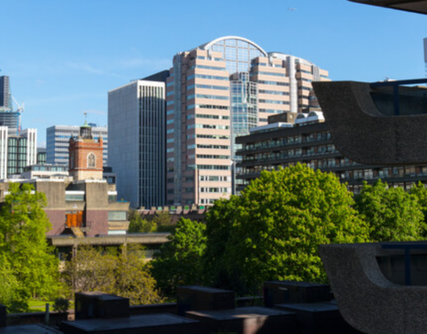 London skyline viewed from the Barbican, with modern office towers, leafy trees, and dark angular Barbican buildings in the foreground.