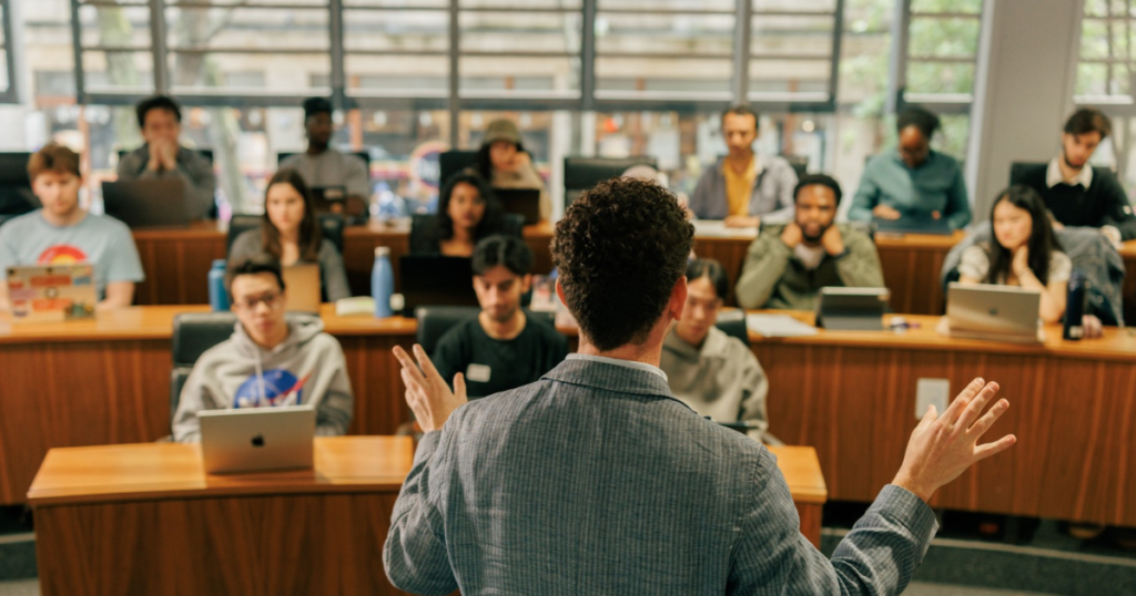 A lecturer stands at the front of a university classroom, seen from behind while addressing a group of students seated in tiered rows. Several students are using laptops, and large windows at the back of the room let in natural light. The classroom has wooden desks and a collaborative learning environment.