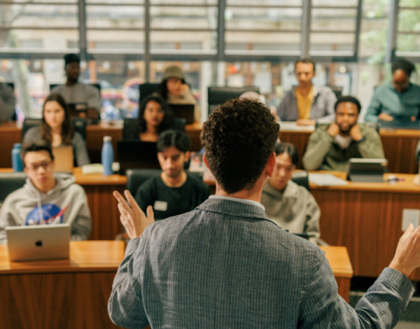 A lecturer stands at the front of a university classroom, seen from behind while addressing a group of students seated in tiered rows. Several students are using laptops, and large windows at the back of the room let in natural light. The classroom has wooden desks and a collaborative learning environment.