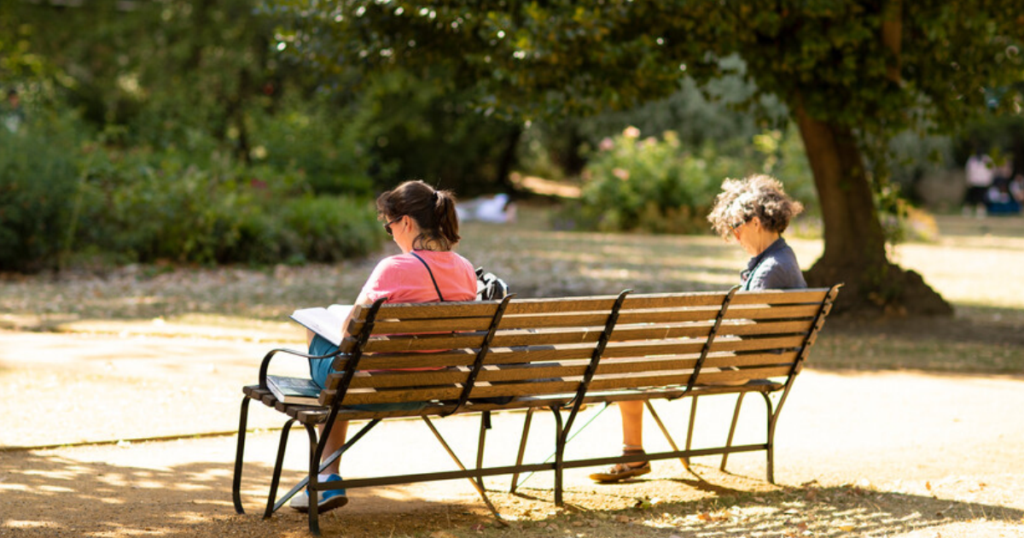 Two people sit on a wooden park bench in a sunny garden; one holds an open book.